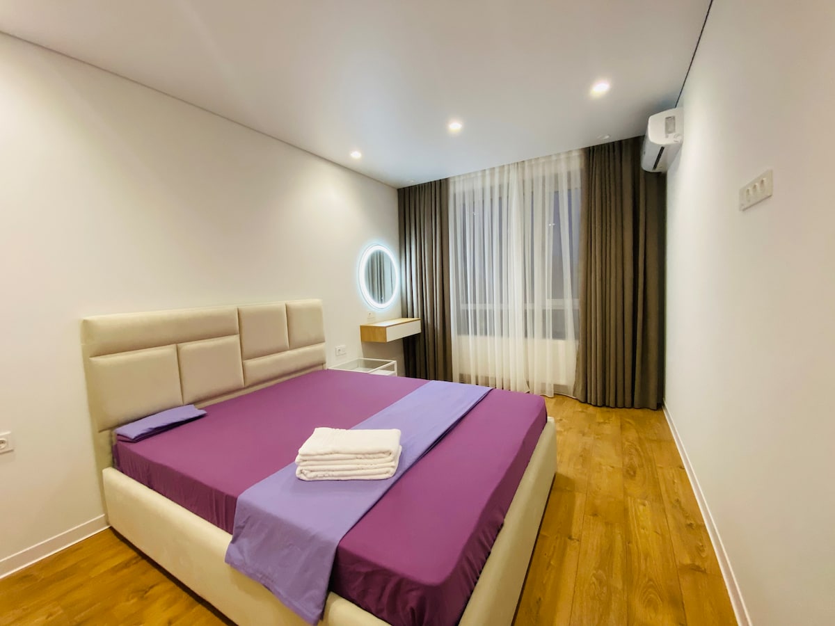 A spacious sleeping area features a large bed with a purple blanket and white linens. A round mirror is positioned near a window, which is covered by sheer curtains that gently diffuse the light. Light-colored walls complement the wooden floor.