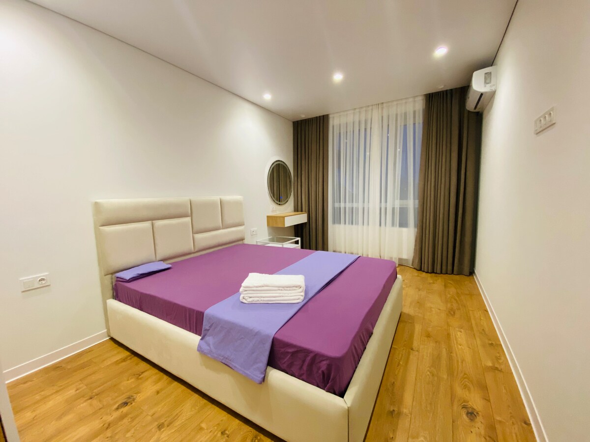 The sleeping area features a spacious bed with a soft headboard, adorned with purple and lavender linens. A circular mirror is mounted above a minimalist desk, while large windows with sheer curtains offer natural light, complemented by warm wood flooring.