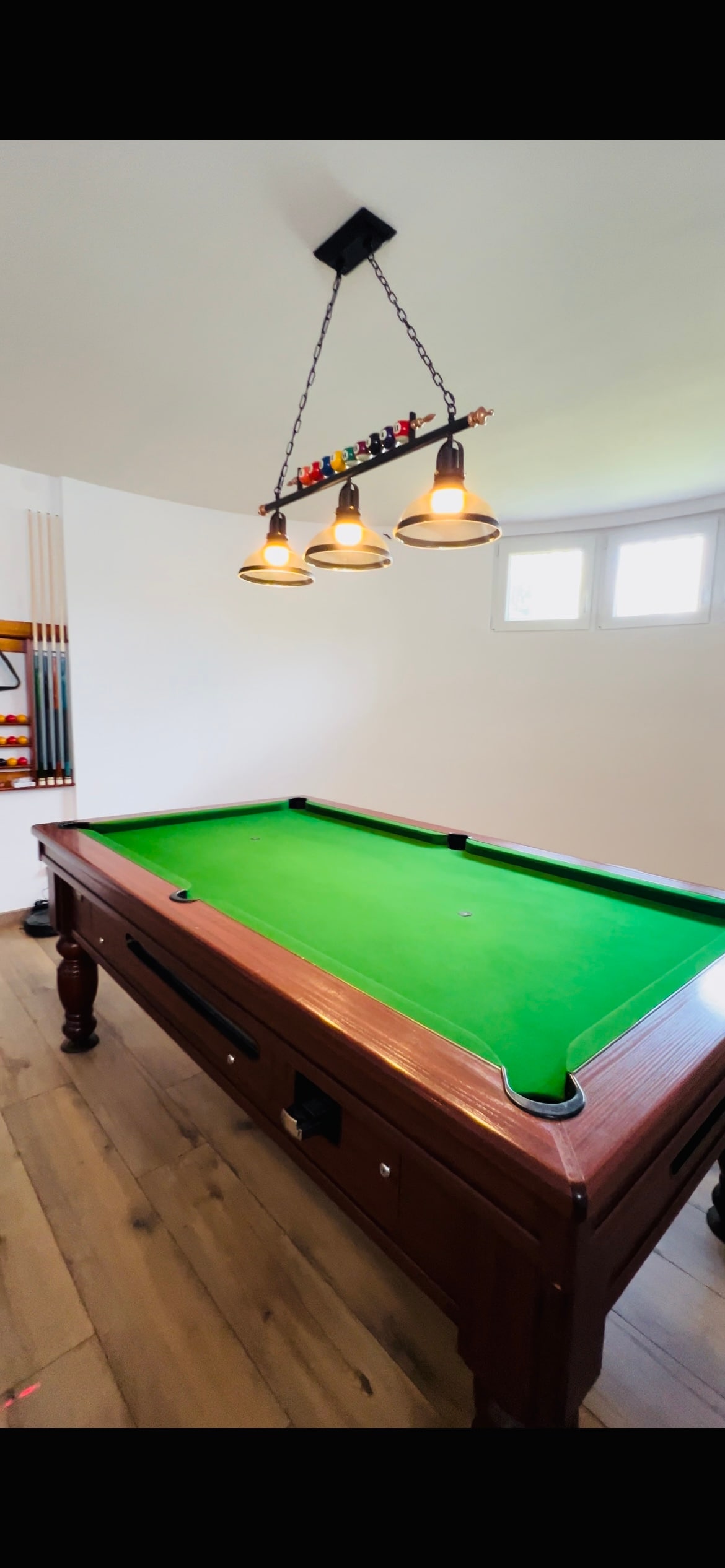 A well-maintained pool table features a vibrant green felt surface, under three pendant lights that provide ample illumination. Two windows are visible, allowing natural light to enter the space, enhancing the overall play area.