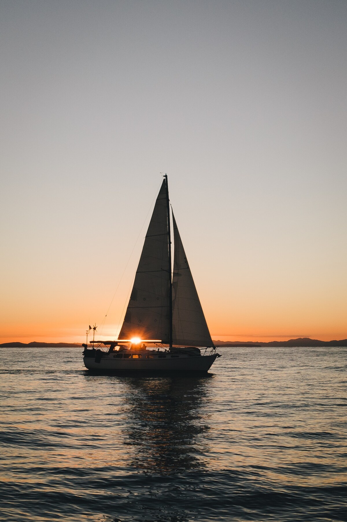 A wooden sailboat is silhouetted against a vibrant sunset, casting reflections on the calm water. The sails are raised, and the warm hues of orange and pink stretch across the horizon, creating a serene and tranquil seascape.