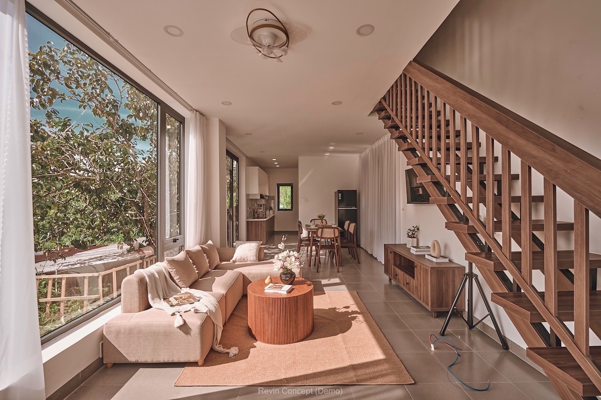 A bright living area features large windows allowing natural light to fill the space. A comfortable sofa surrounds a round wooden coffee table, with a soft area rug beneath. A staircase with wooden railings leads to the upper level, enhancing the open layout.