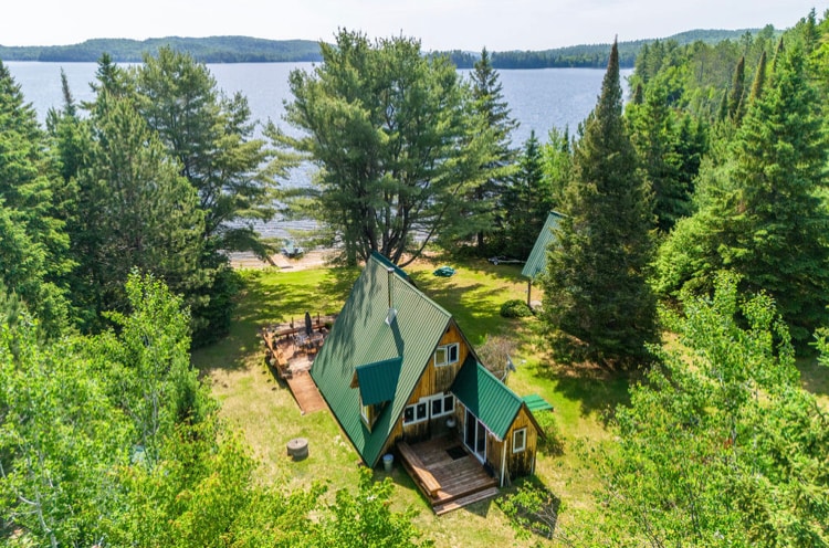 A cabin with a green metal roof is surrounded by trees, situated on the shoreline of Aylen Lake. Clear views of the water are visible from the property, and a sandy beach is nearby. An outdoor deck offers space for relaxation.