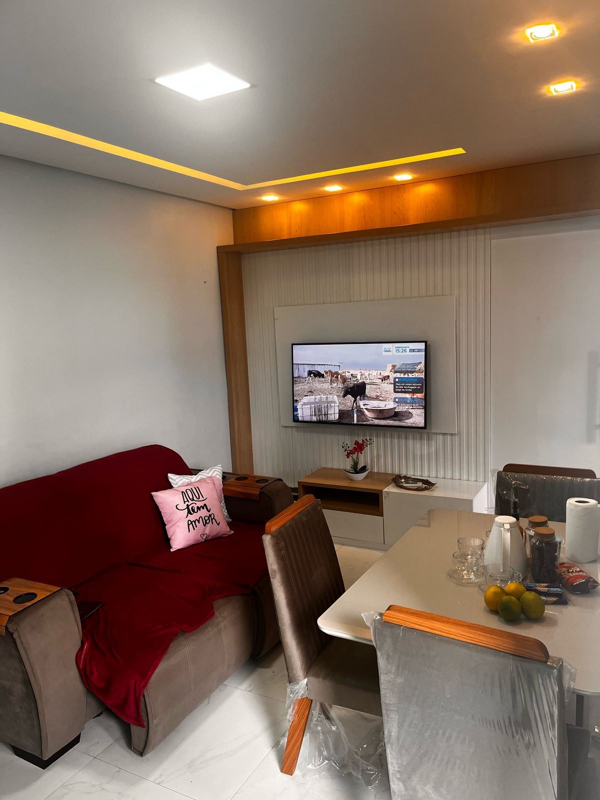 A comfortable living area is presented with a soft sofa covered in a maroon throw. A television is mounted on the wall, accompanied by a dining table set for two. Natural light filters in, highlighting the warm tones of the decorative elements.