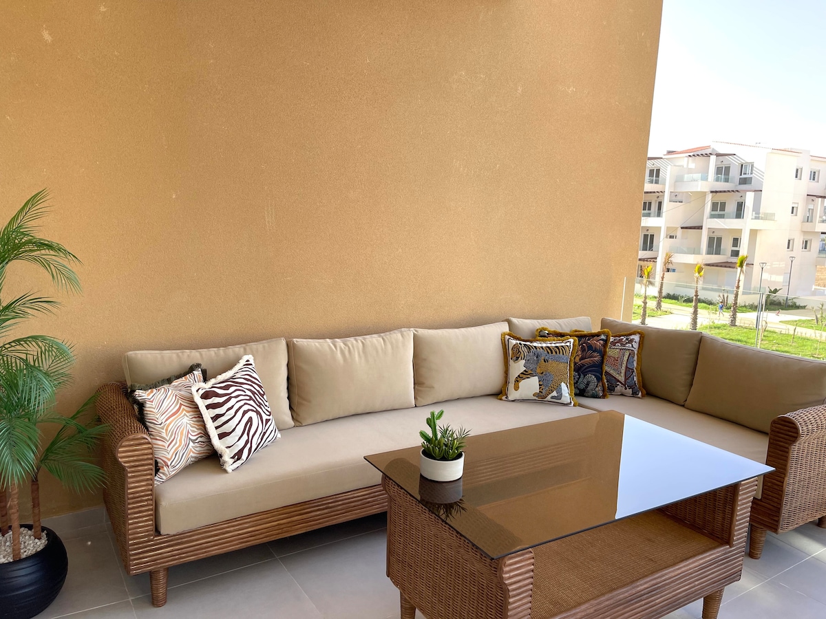 A stylish outdoor seating area is presented, featuring a spacious sectional sofa adorned with patterned cushions. A sleek glass-top table is positioned at the center, complemented by a small potted plant. The warm, textured wall provides a backdrop to this inviting space.