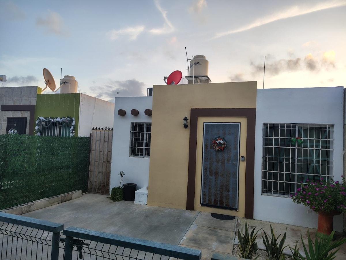 A modest exterior of a single-story house is shown, featuring a welcoming front door adorned with a seasonal wreath. Two satellite dishes are visible on the roof. A small garden with green foliage and potted plants frames the entrance, creating an inviting appearance.