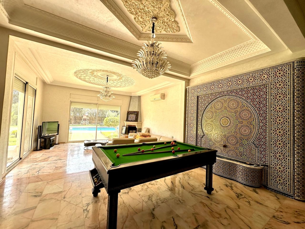 An elegantly decorated game room features a stylish pool table, surrounded by intricately designed walls and a decorative ceiling. Large windows allow natural light to fill the space, while showcasing views of the outdoor pool area.