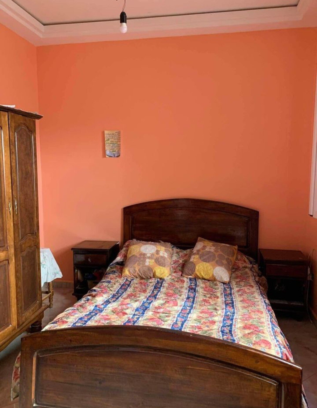 A warm-toned bedroom features an inviting wooden bed adorned with a floral bedspread and two pillows. Flanking the bed are two nightstands, and a wooden wardrobe is positioned against the orange walls, complemented by a simple piece of wall art.