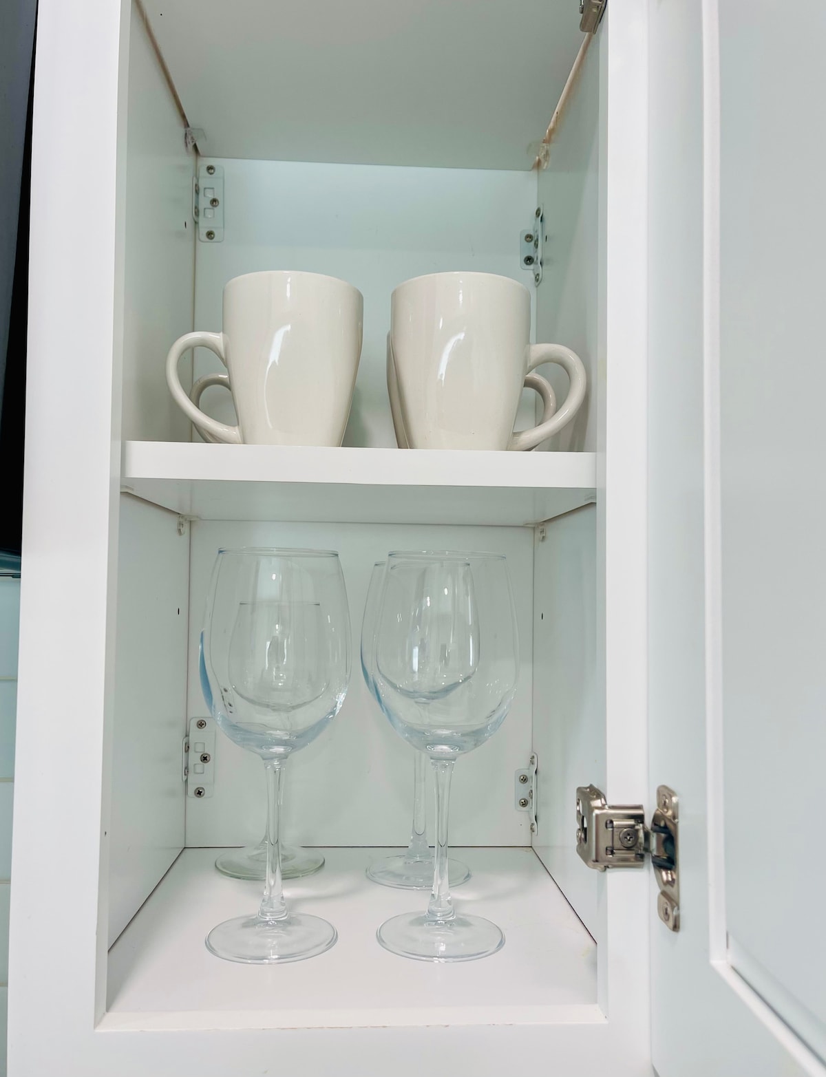 A clean white cabinet contains two matching ceramic mugs positioned above two elegant glass wine glasses. The mugs are of a simple design, while the wine glasses feature a tall stem. The interior of the cabinet is bright, highlighting its organized storage.