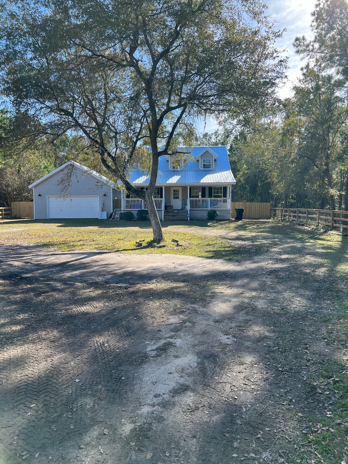 A two-story farmhouse is seen nestled among trees, featuring a blue metal roof and a spacious front porch. A gravel driveway leads to the house, which is surrounded by a large, grassy yard. A wooden fence encloses the property, enhancing the serene outdoor surroundings.