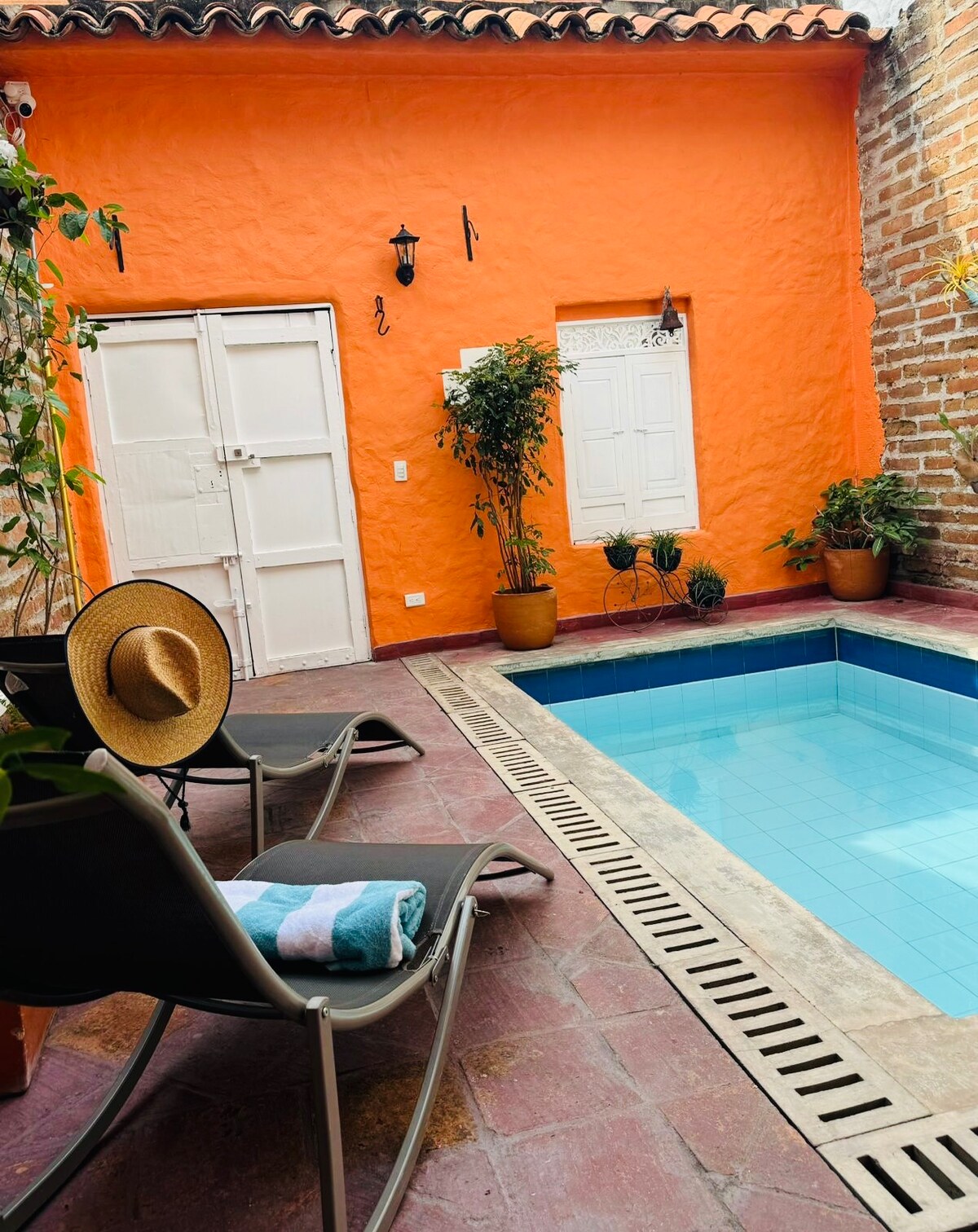 A vibrant orange wall frames a small outdoor area featuring a refreshing pool. Two lounge chairs with towels are arranged beside the pool. Potted plants add greenery to the space, while a white door and window provide brightness and charm.