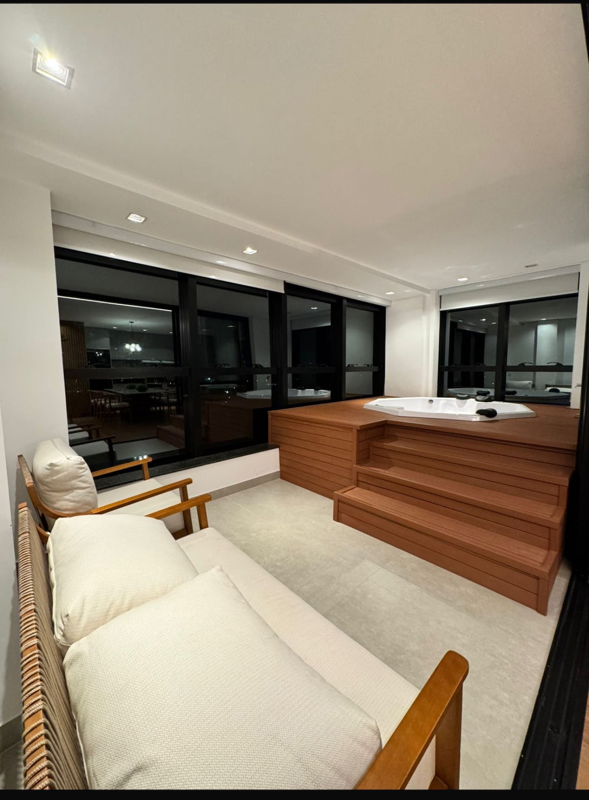 A well-lit relaxation area is presented, featuring a wooden jacuzzi positioned against a backdrop of large windows. A comfortable seating arrangement is provided nearby, subtly complementing the spa-like atmosphere. Soft lighting enhances the serene environment, inviting restful moments with a view of the city.