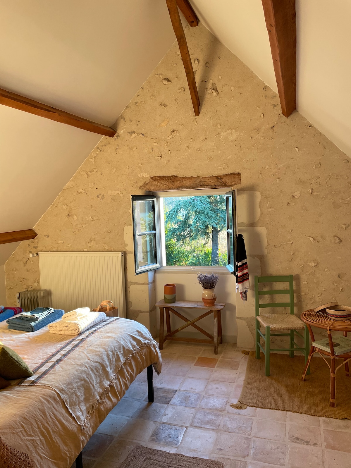 An inviting bedroom features a spacious bed dressed in neutral linens. A large window offers views of the greenery outside, while natural light floods the room. Wooden beams accentuate the rustic ambiance, complemented by a small table and a chair made from woven materials.