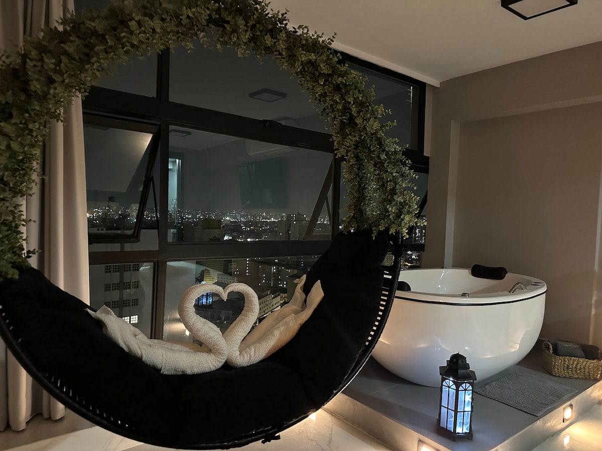 A cozy seating area features a round, decorated swing chair beside a modern bathtub. The large windows offer a stunning nighttime view of the city skyline, while soft lighting creates an inviting ambiance.
