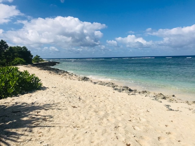 A sandy beach is visible, bordered by lush green foliage and rocky outcrops. The calm blue waters stretch into the horizon, with gentle waves lapping at the shore. A clear sky with fluffy white clouds enhances the serene coastal landscape.