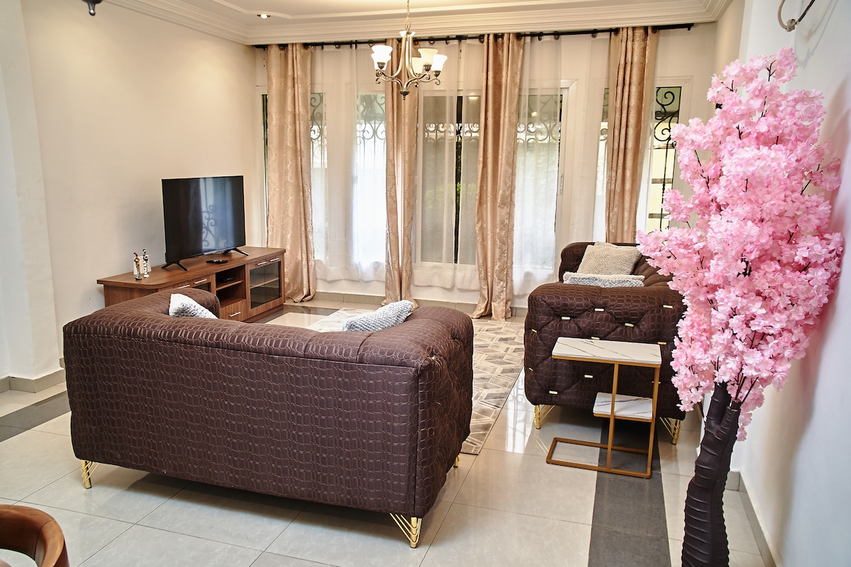 A living area is presented with two brown sofas arranged for comfort. A TV sits on a wooden console against a wall, while a small side table stands next to one sofa. Natural light filters through large windows, framed by sheer curtains, with a decorative pink blossom tree adding a touch of color.