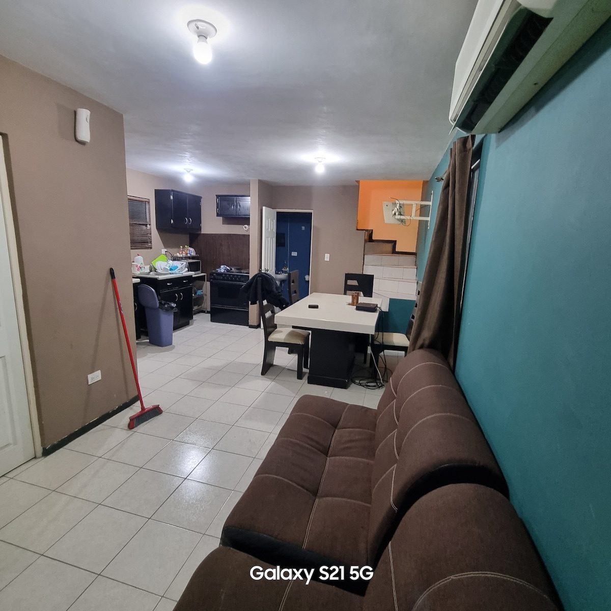 An open living space features a comfortable sofa facing a dining table. The kitchen, visible in the background, includes dark cabinetry and appliances. Brightly colored walls add dimension. Natural light enters through a window, and an air conditioning unit is mounted above.