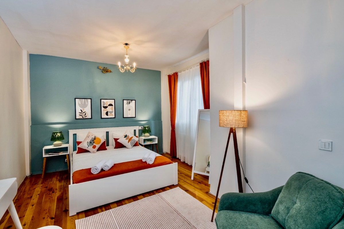 A well-lit bedroom features a spacious bed with white linens and decorative pillows. Framed artwork adorns the wall, complemented by two nightstands with lamps. A cozy green armchair sits beside a stylish floor lamp, enhancing the inviting ambiance of the room.