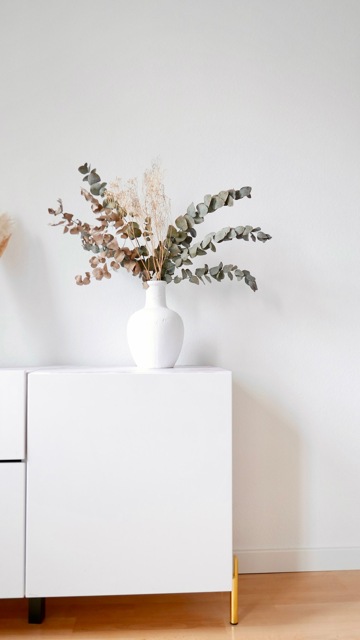 A white cabinet is positioned against a light wall, topped with a simple white vase. The vase holds a mix of dried foliage, including eucalyptus and small, delicate stems, adding a touch of natural decor to the space.