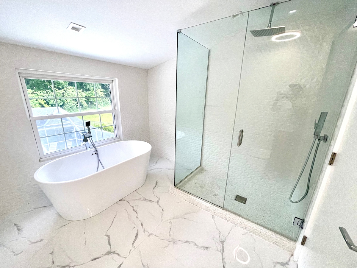 A spa-like bathroom is showcased, featuring a freestanding tub beside a glass-enclosed shower. Natural light enters through a large window, illuminating the elegant marble flooring and textured wall. Fixtures are sleek and modern, creating a calming space for relaxation.