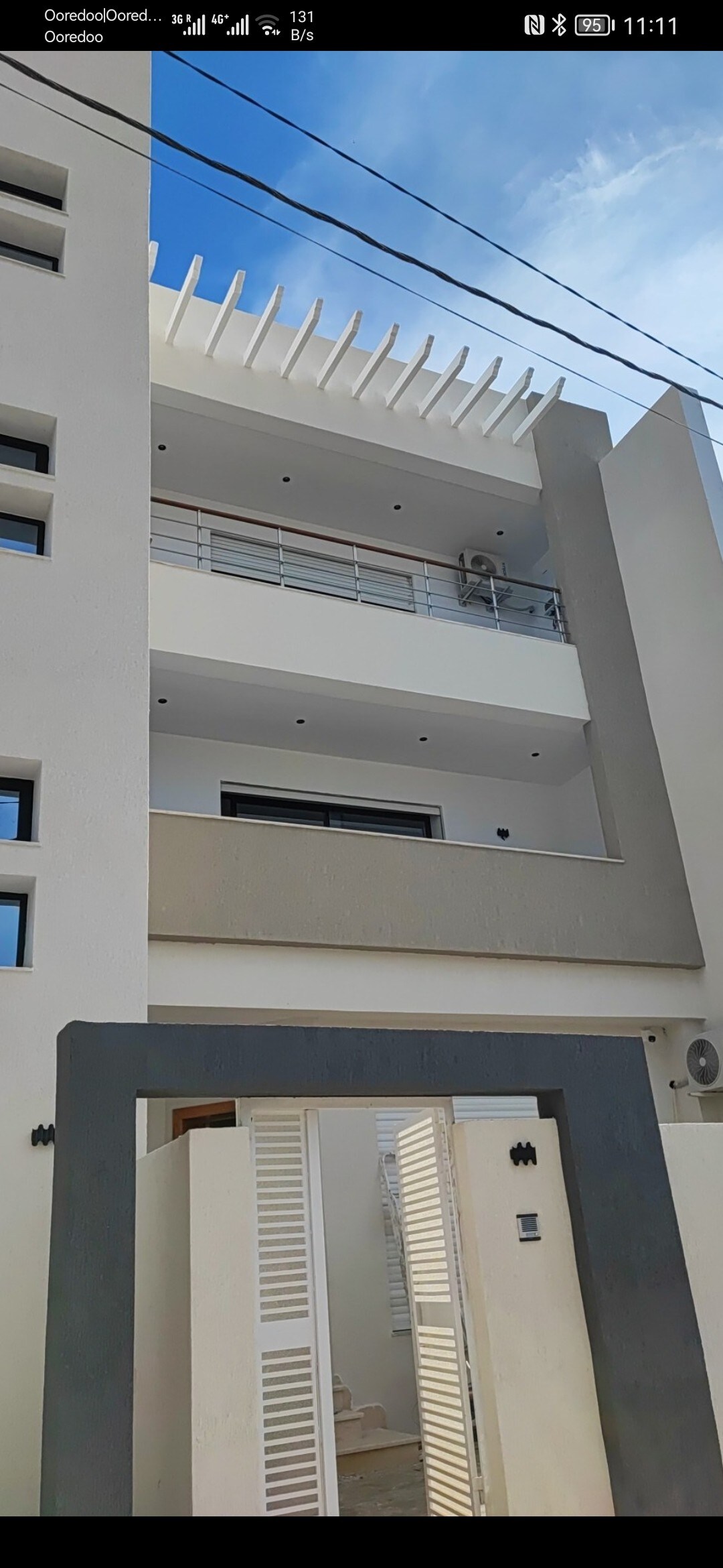 A modern building exterior is shown, featuring a two-story façade with a balanced mix of clean lines and neutral tones. A spacious balcony is visible on the second floor, framed by sleek railings. The entrance is marked by a minimalist gate and decorative elements.