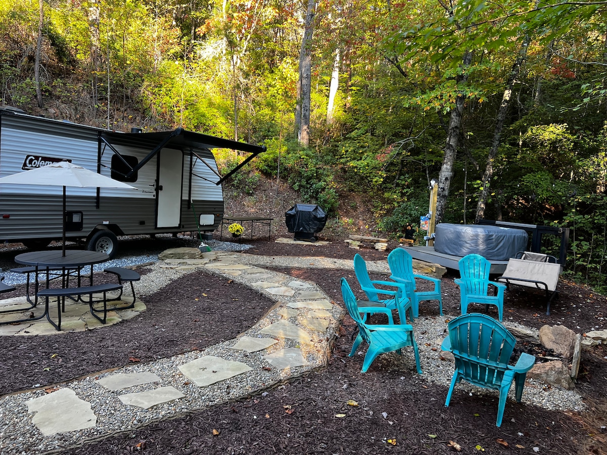 An outdoor space features a gravel path lined with natural stones, leading to a seating area with several blue chairs. A picnic table sits nearby, with a grill and hot tub visible in the background, surrounded by trees showcasing vibrant autumn colors.