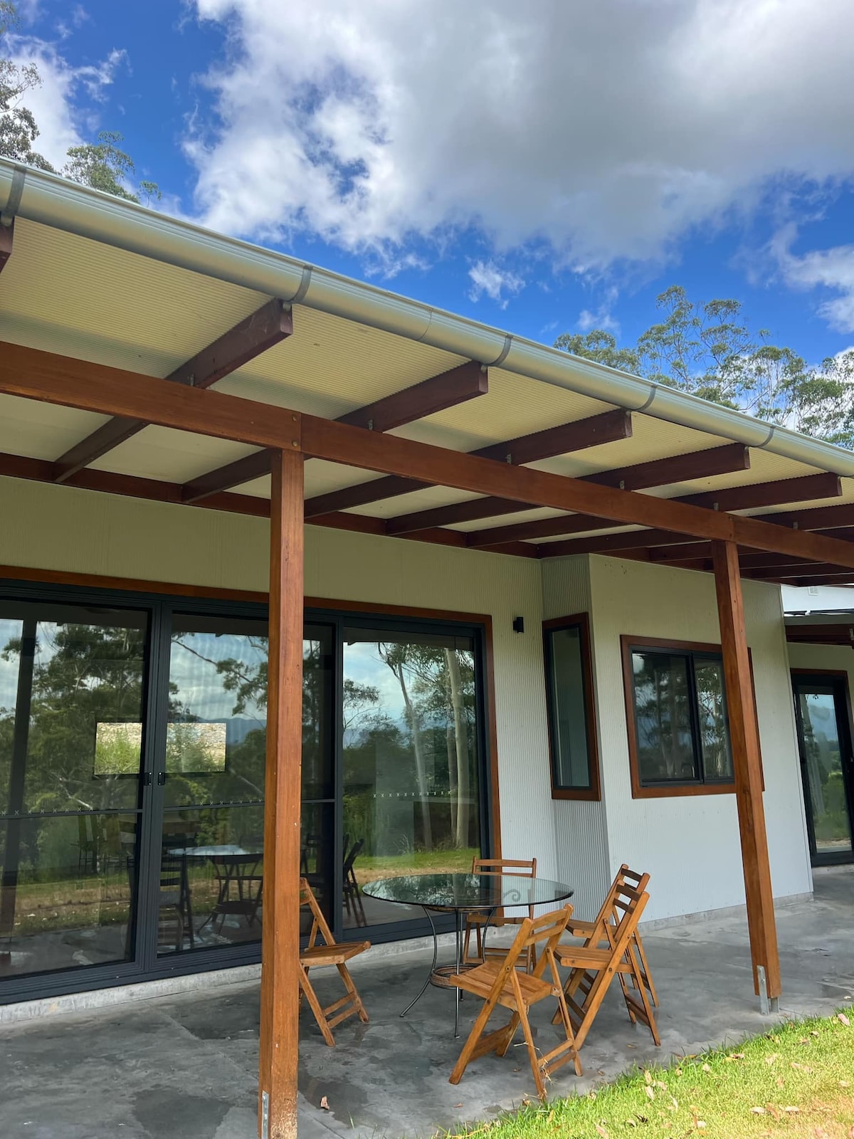 A covered outdoor seating area is featured, showcasing a glass table surrounded by four wooden chairs. Large sliding glass doors provide access to the interior, with greenery visible through the windows and a partially cloudy sky above.