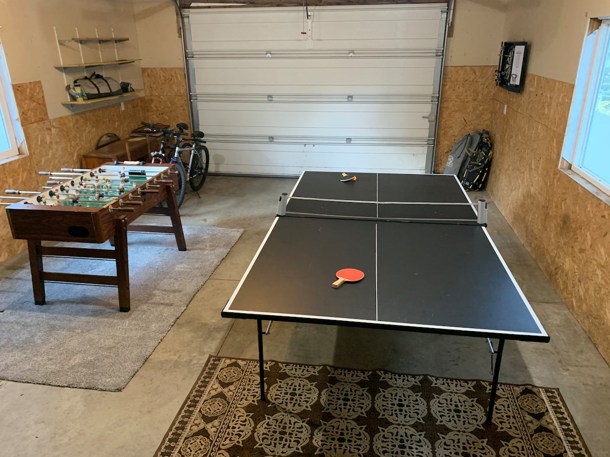 An indoor recreational space features a ping pong table and a foosball table, both ready for play. The concrete floor is covered by a textured area rug, and natural light enters through two large windows, creating a bright and open atmosphere.