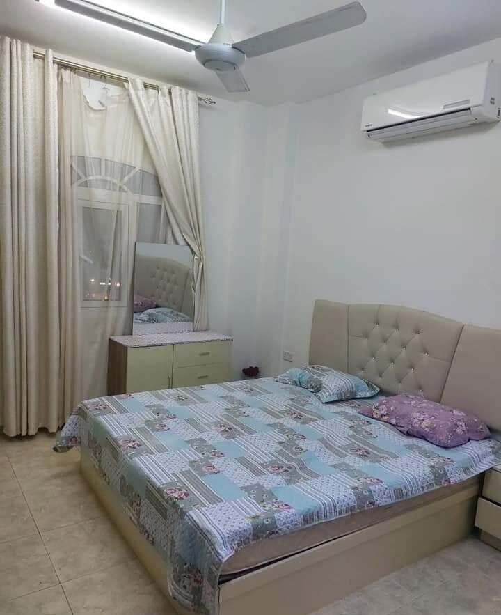 A light-filled sleeping area features a spacious bed dressed in a patterned blanket. Soft pillows add comfort. A mirrored dresser is positioned nearby, complementing the room's clean design. Floor-to-ceiling curtains frame the window, allowing natural light to enter the space.