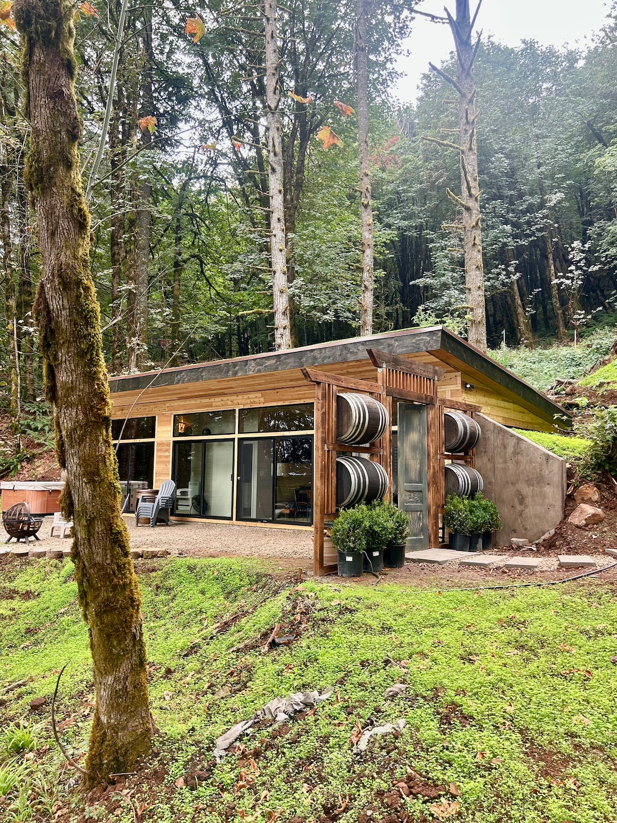 The exterior of the unique home features a streamlined design surrounded by lush greenery. Sleek glass walls offer visibility into the interior, while large wine barrels are placed against the structure, enhancing its rustic charm. A gravel pathway leads up to a cozy seating area.