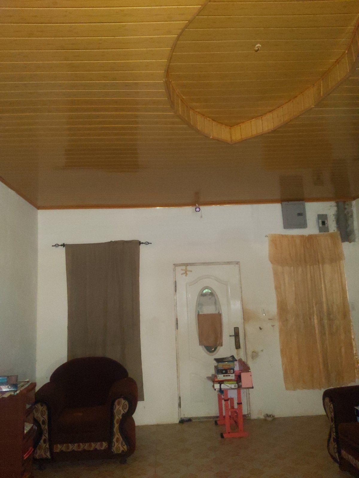 A living area showcases a golden wood-paneled ceiling reflecting light. One wall features a door with an oval mirror and two windows dressed with beige curtains. A maroon armchair is positioned near a small pink table, complemented by wooden furniture in the background.