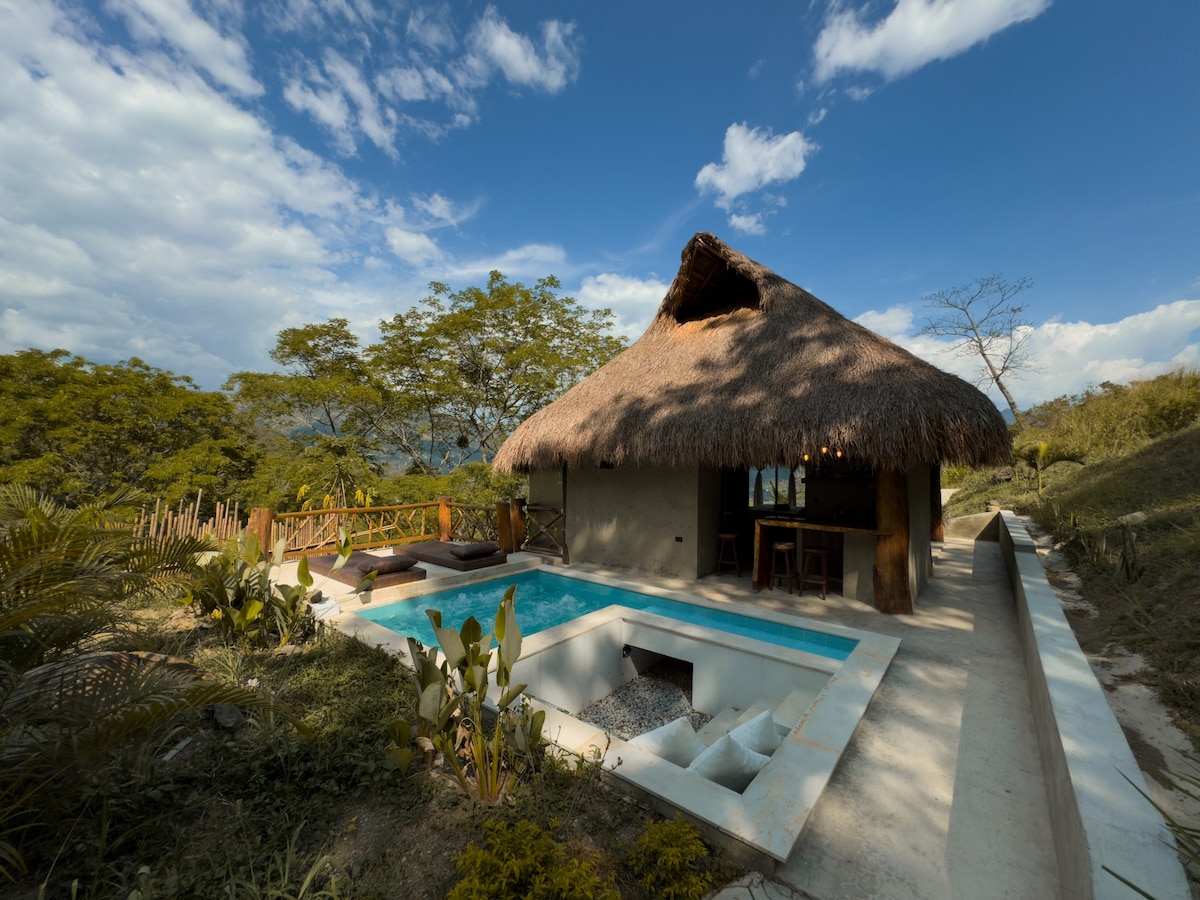 A traditional thatched-roof structure is viewed alongside a private pool, surrounded by lush greenery. The outdoor space features a patio area with seating options. The natural landscape includes distant mountains and a clear blue sky, enhancing the tranquil atmosphere.