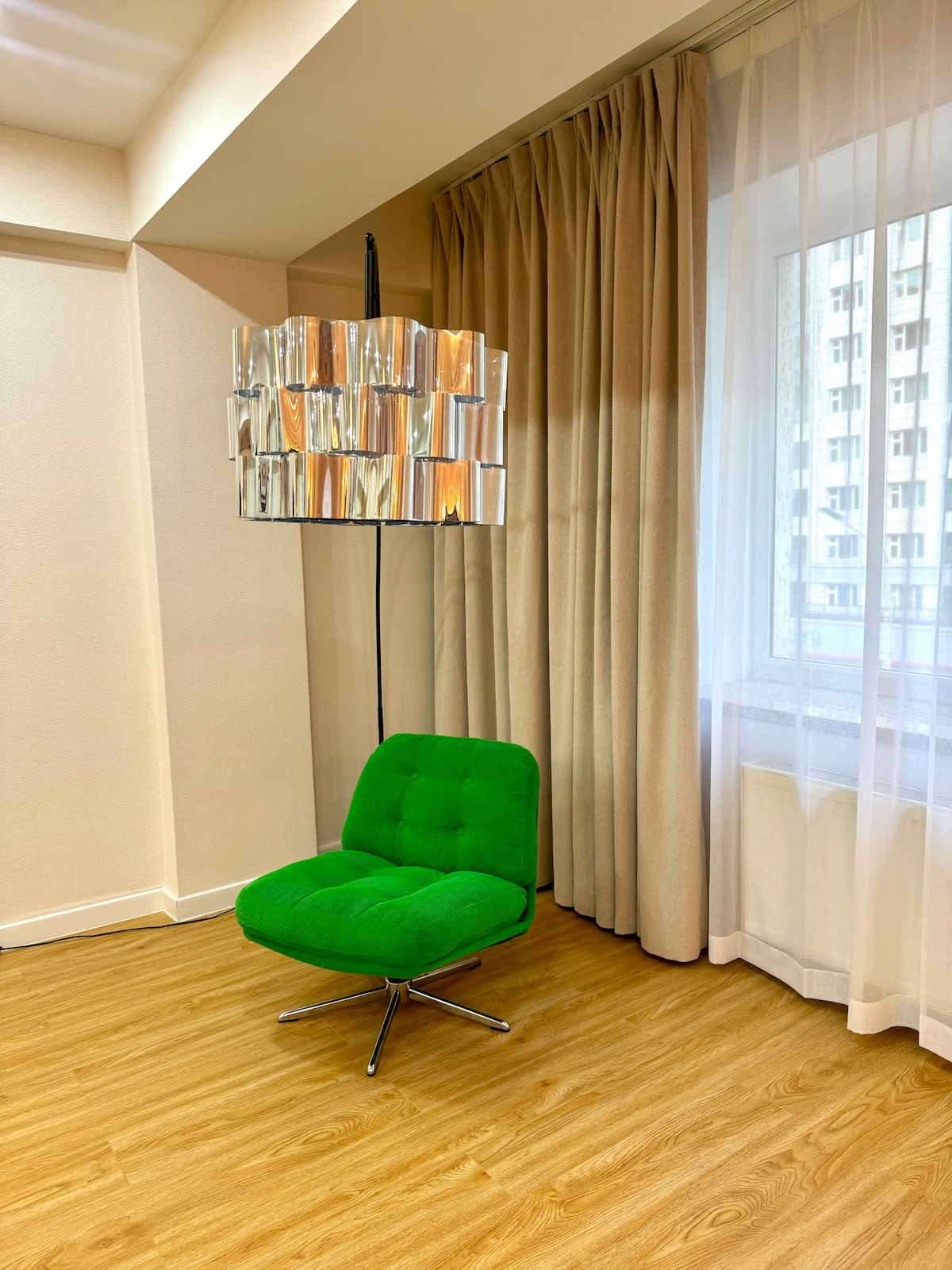 A modern green chair with a tufted design is positioned near a large window, allowing natural light to illuminate the space. Above, a contemporary light fixture with reflective elements adds a stylish touch. Soft, beige curtains frame the window, complementing the warm wood flooring.