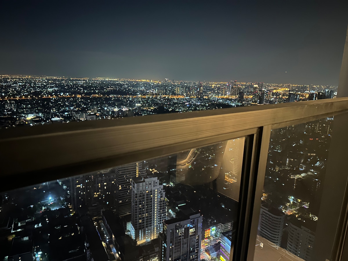 A panoramic view showcases the vibrant city lights of Bangkok at night, framed by large glass windows. A distant skyline is visible, with buildings illuminated under a dark sky, creating a stunning contrast against the urban landscape.