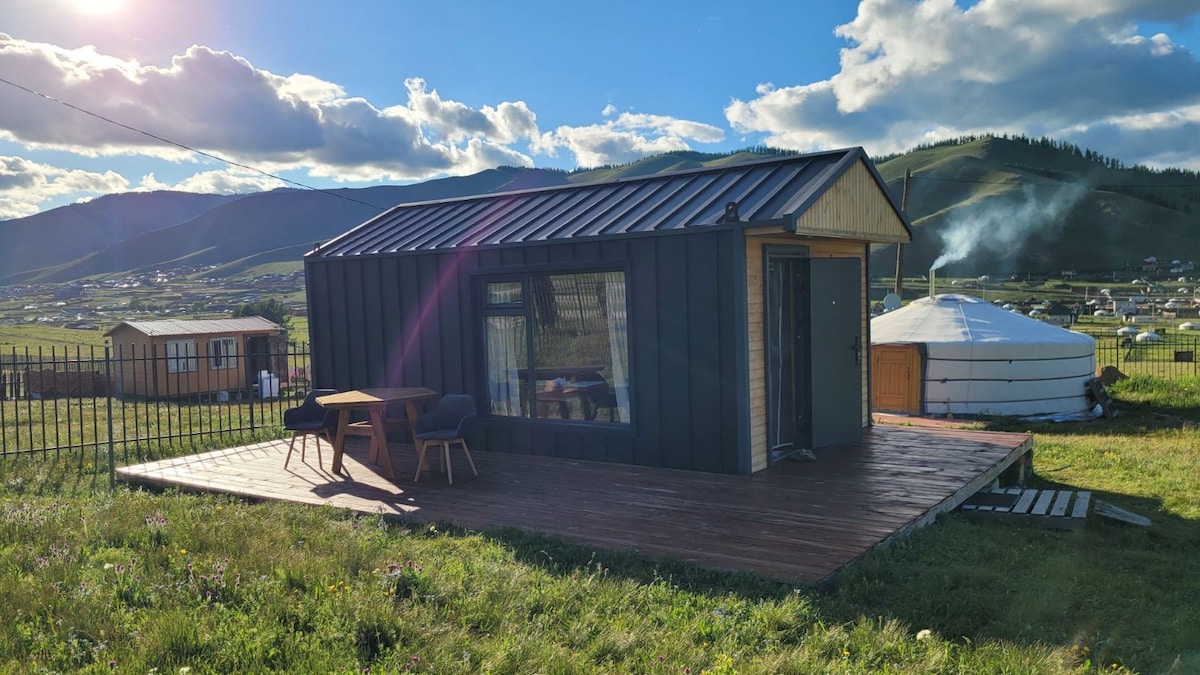 A tiny house with a metal roof is positioned on a wooden deck, surrounded by lush green fields and distant mountains. The side of the house features large glass windows, allowing natural light to fill the interior. Nearby, a traditional yurt is visible in the background.