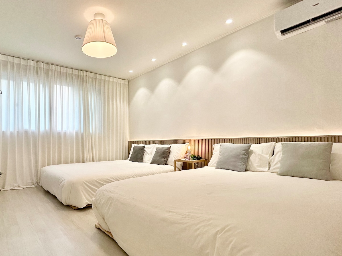 A bright bedroom features two spacious beds with soft bedding, complemented by decorative pillows. A soft, natural light filters through sheer curtains, adding warmth to the white walls. A small bedside table with a lamp enhances functionality in the room.