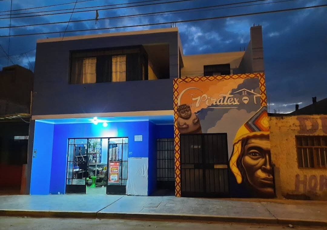 The exterior of a two-story building is shown, featuring a vibrant blue façade with a mural on one side. Large glass windows provide a view into the interior, which is visible through the open entrance. The scene is illuminated by a warm light, suggesting evening hours.