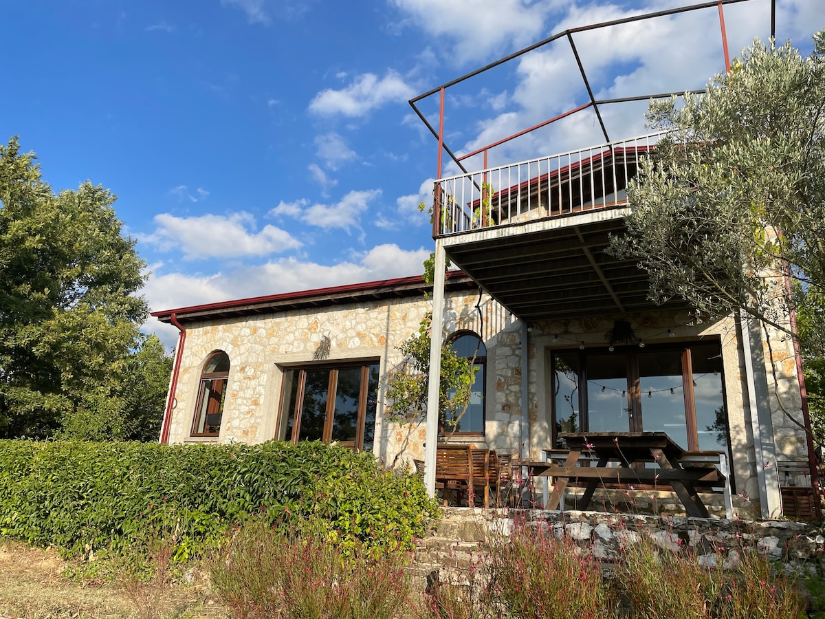 A charming stone house with large windows is surrounded by lush greenery. A covered terrace offers outdoor seating, while a picnic table is positioned near the entrance. The blue sky and white clouds create a serene atmosphere above the property.