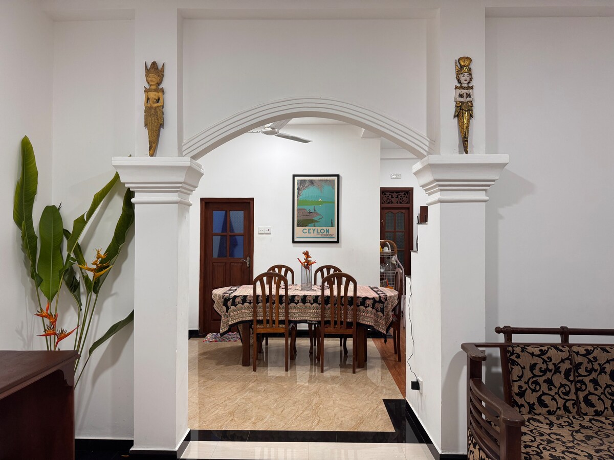 An archway leads into a dining area featuring a large table surrounded by wooden chairs. Artwork depicting Ceylon is displayed on the wall. Decorative statues frame the entrance, and greenery is visible in the corner, adding a touch of vibrancy to the space.