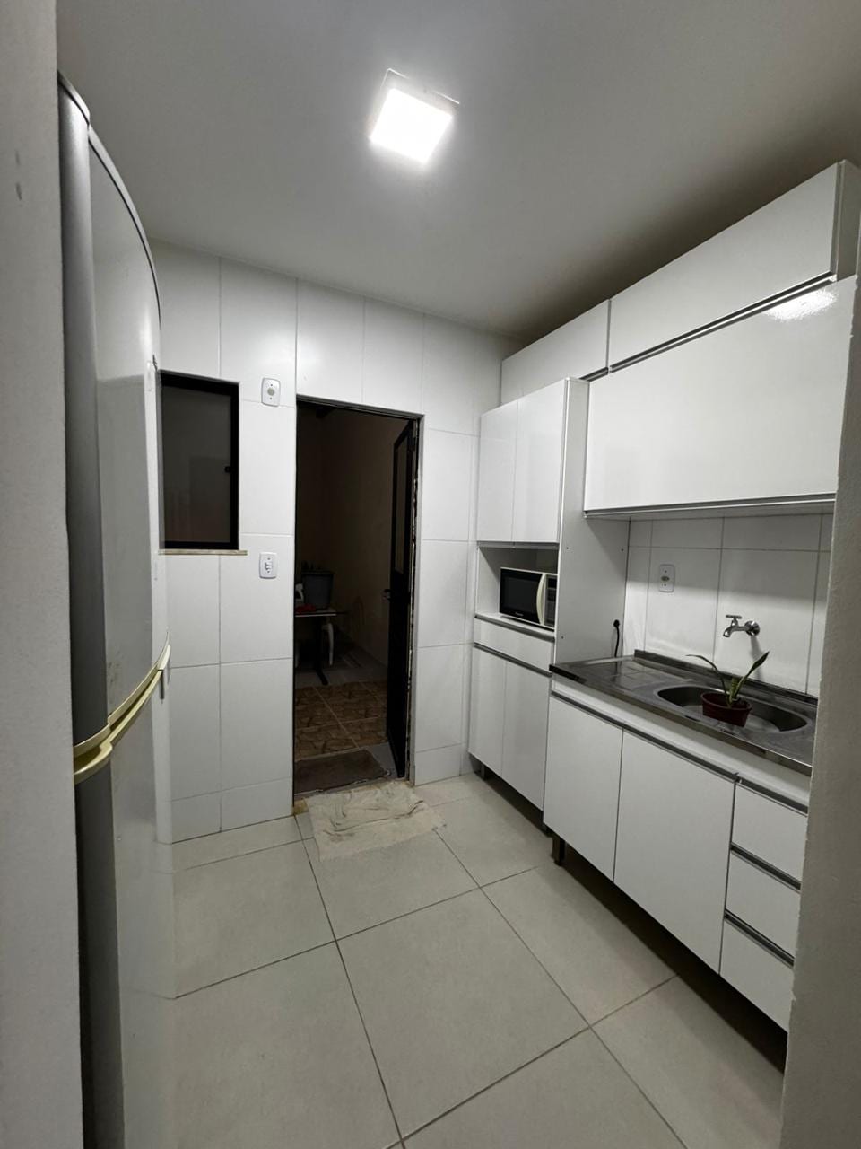 A modern kitchen features sleek white cabinetry and a stainless steel refrigerator. Natural light enters through a window above the sink, where a small plant is placed. The clean tiles and minimalist design create a functional and welcoming cooking space.