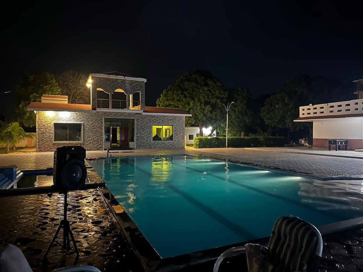 A spacious pool area is illuminated at night, featuring a large swimming pool reflecting soft lights from the surrounding buildings. A two-story structure can be seen, with large windows offering glimpses of the interior. Lush trees line the background, enhancing the natural ambiance.