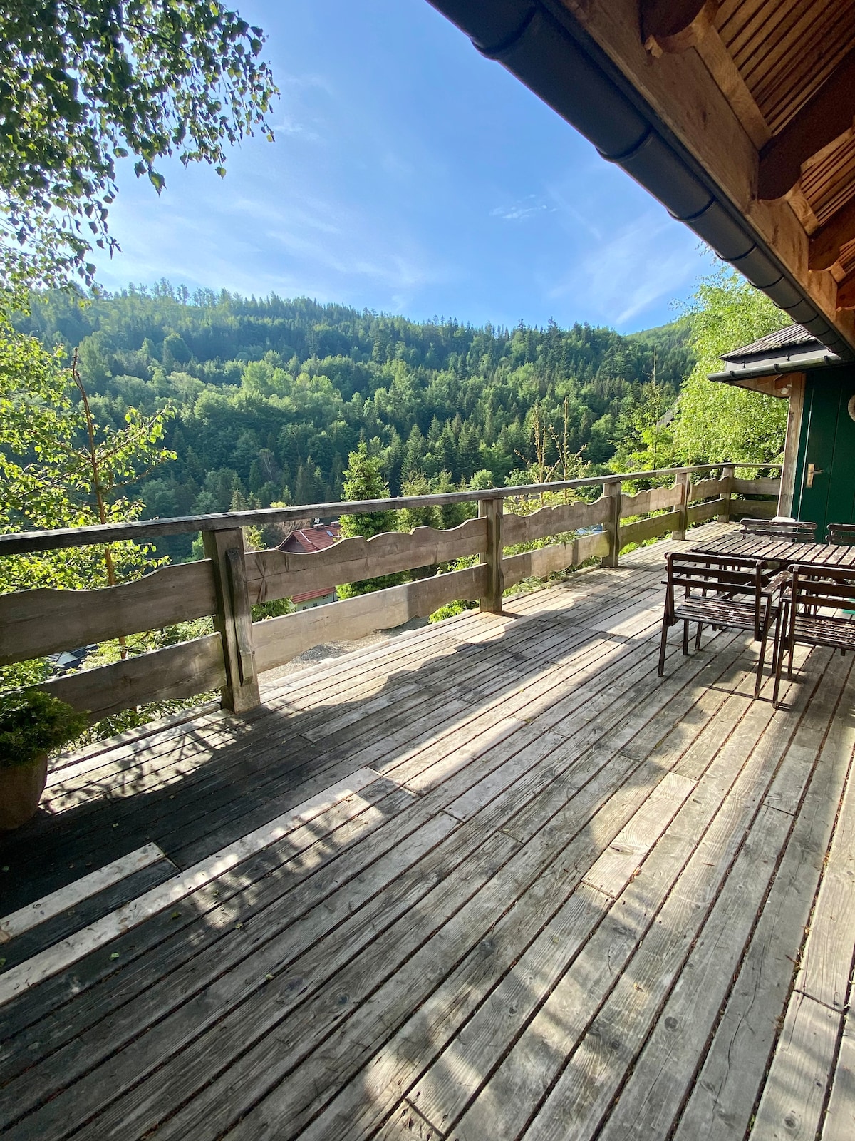 An expansive wooden terrace offers unobstructed views of the surrounding green hills and forests. A set of outdoor furniture, including a table and chairs, is positioned on the deck, inviting relaxation and enjoyment of the natural scenery.