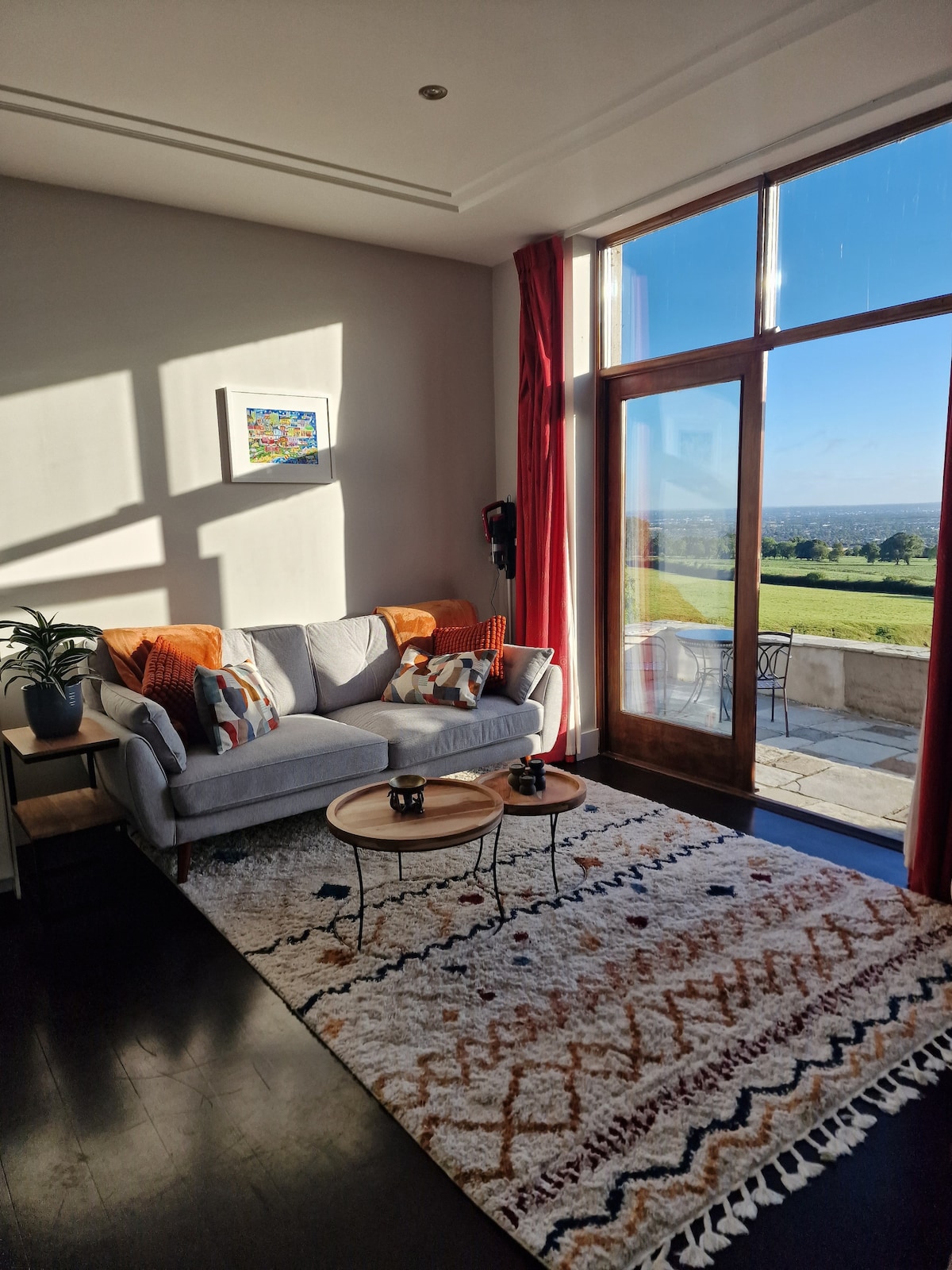 A bright living area is showcased, featuring a comfortable sofa adorned with patterned cushions. Two small tables sit in front, and a large glass door opens to reveal picturesque views of the outdoor landscape. Natural light filters in, casting gentle shadows across the room.