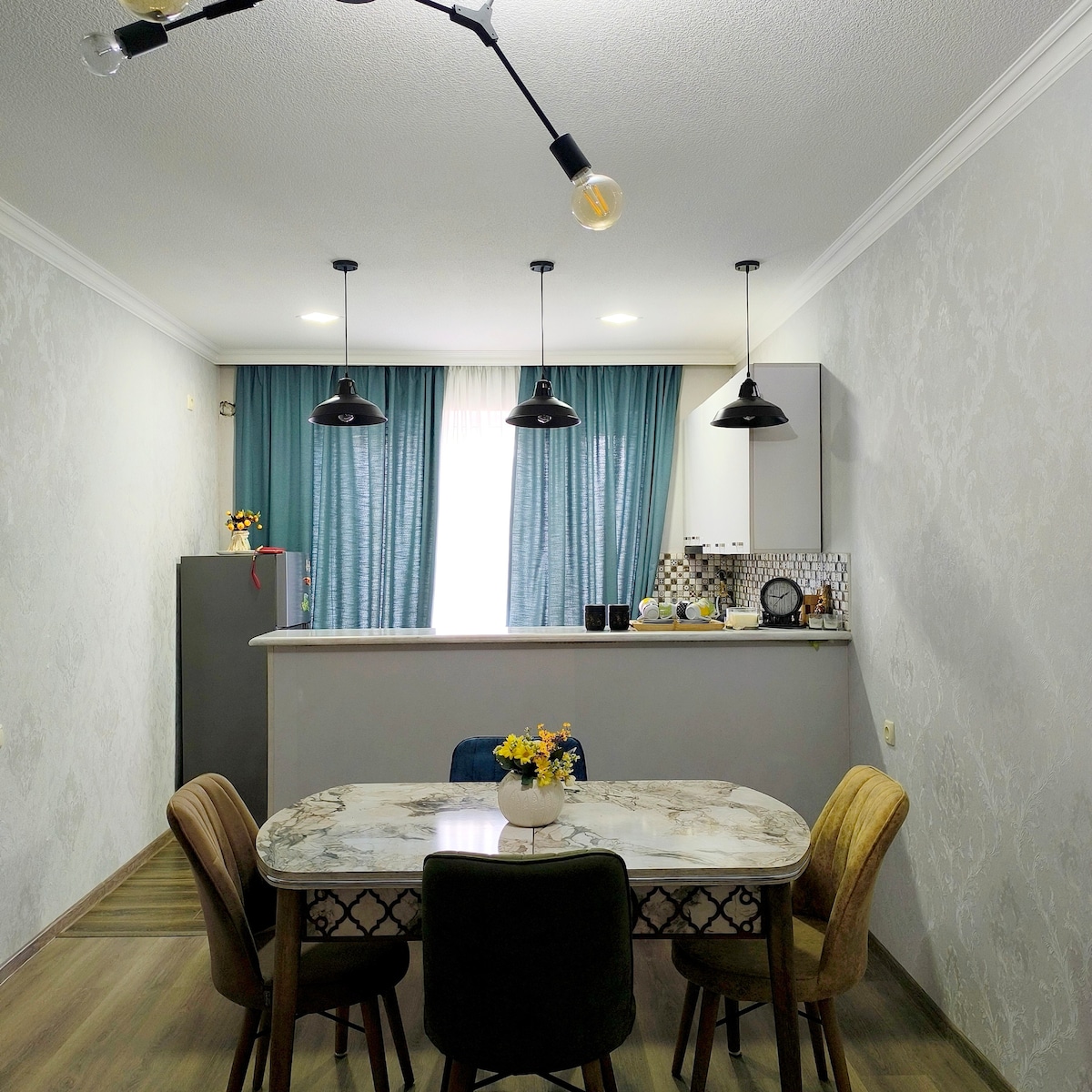 A dining area is shown with a marble-top table surrounded by four upholstered chairs in varied colors. Overhead, three pendant lights provide illumination. In the background, a kitchen equipped with a refrigerator and cabinetry is partially visible, complemented by soft curtains that diffuse natural light.