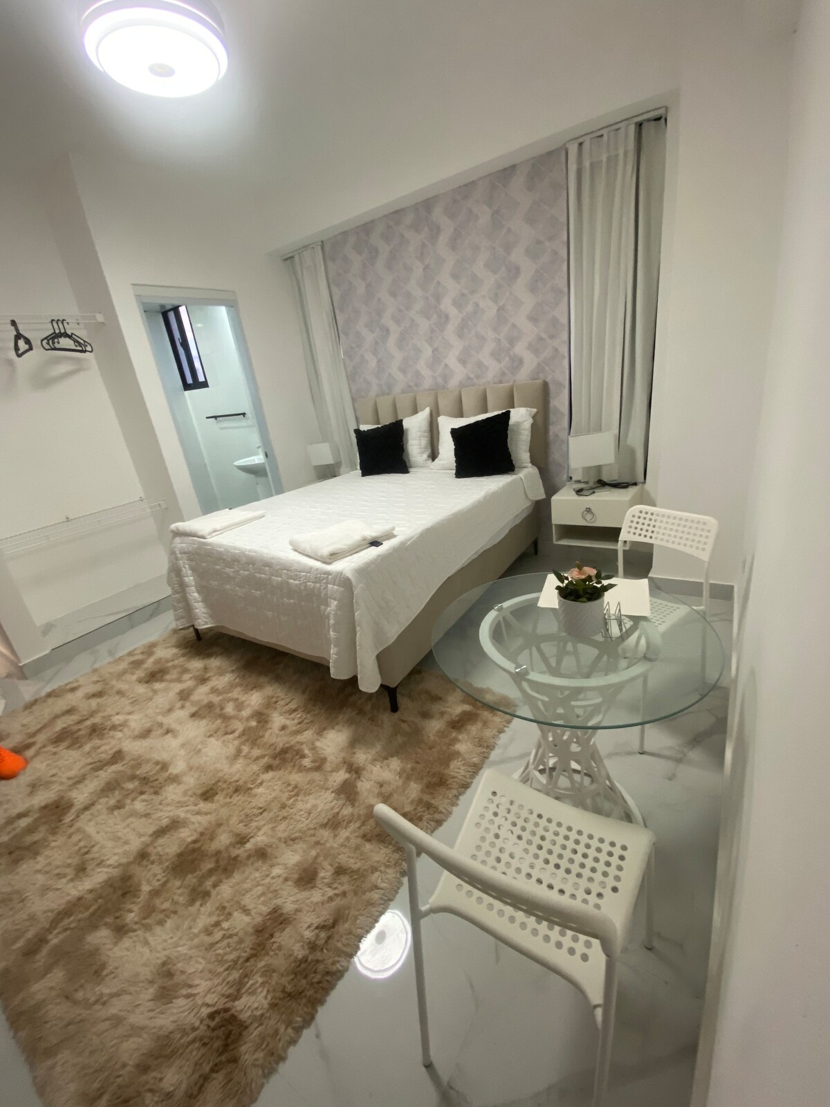 A well-furnished living space is displayed, featuring a spacious bed with neatly arranged pillows. A round glass table sits centrally, surrounded by four white chairs. A soft area rug adds warmth, while a small plant decorates the table. The bathroom entrance is visible on one side.