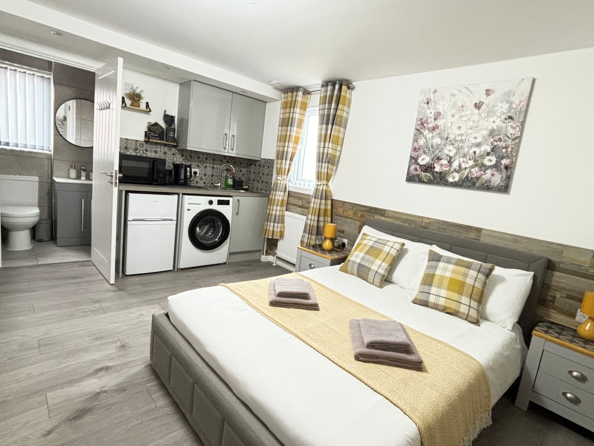 A cozy sleeping area features a spacious bed adorned with folded towels and a yellow blanket. The kitchen area is equipped with modern appliances, while the decor includes light wood accents and a floral artwork on the wall. Natural light flows through the window dressed with plaid curtains.