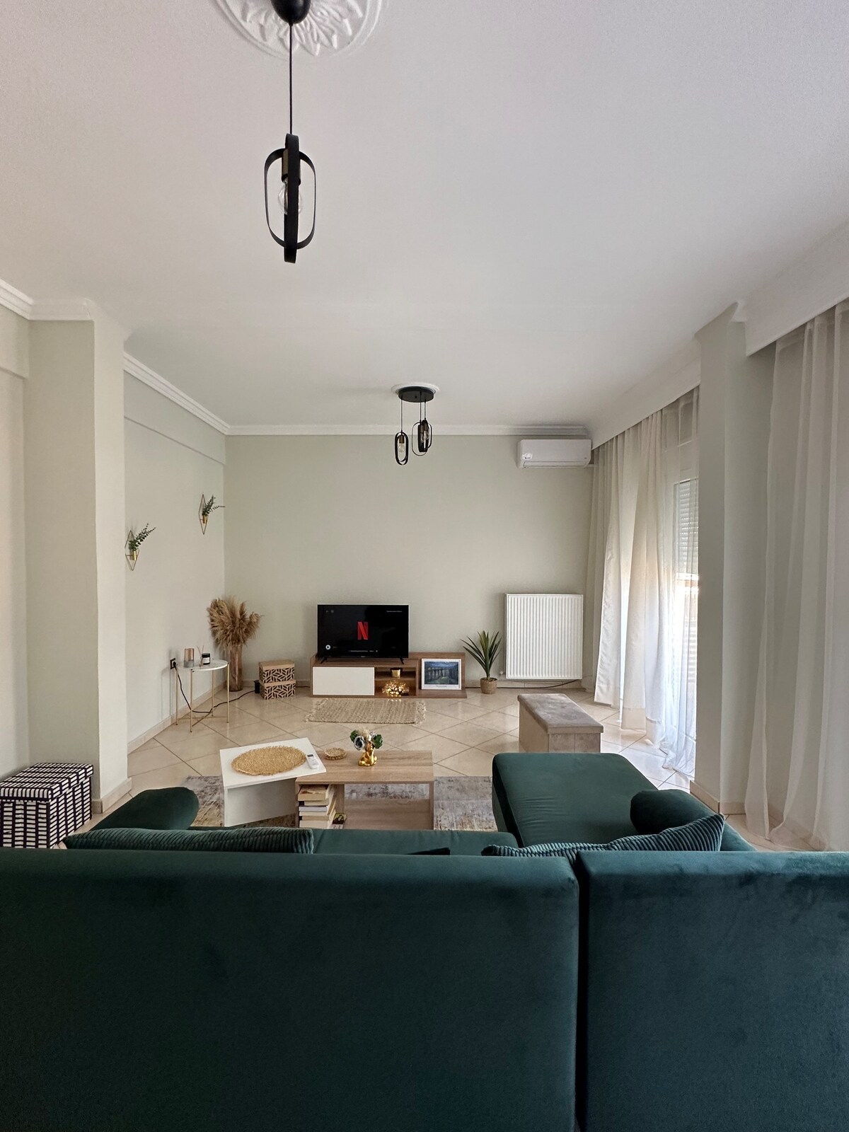 A spacious living area is furnished with a comfortable green sofa facing a sleek television. Natural light filters in through sheer curtains, illuminating the neutral walls and tiled floor. Decorative elements include a small coffee table and wall accents, creating a cozy environment.