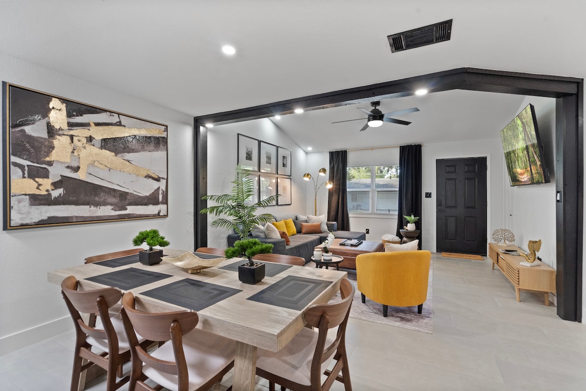 A spacious living and dining area showcases modern decor, featuring a large dining table surrounded by wooden chairs. A plush sectional sofa with vibrant yellow accent chairs invites relaxation. Artwork adorns the walls, and a ceiling fan adds comfort, complemented by natural light from the windows.