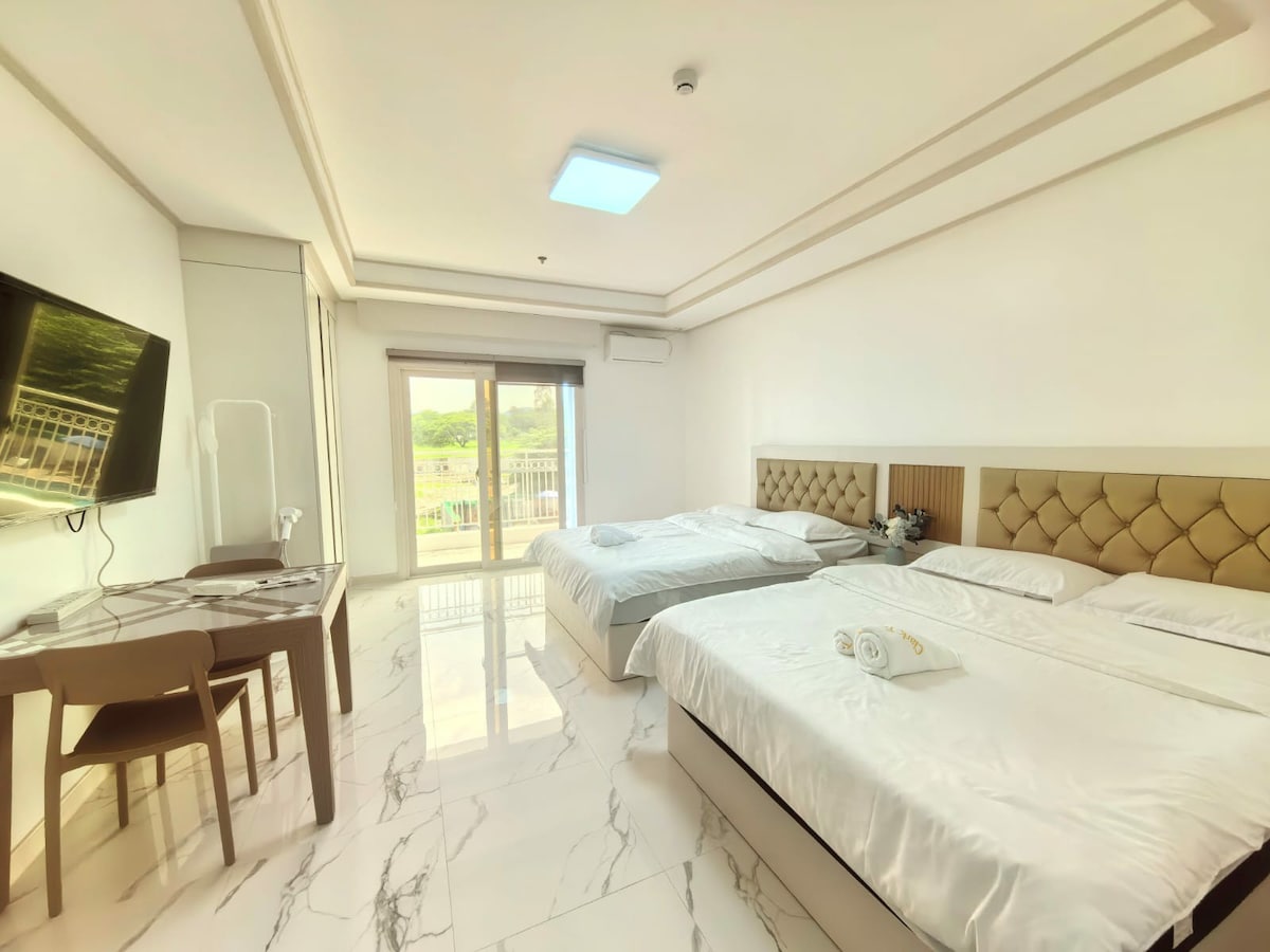 A spacious studio room is presented with two large beds and a modern desk. A flat-screen television is mounted on the wall. Large glass doors provide natural light and access to the balcony, showcasing views of the surrounding area. The marble flooring adds a contemporary touch.