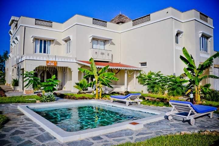 The two-story villa features a private pool surrounded by lounge chairs. Lush greenery and banana plants enhance the outdoor space, while a stone pathway leads to the entrance. The light-colored exterior reflects sunlight against a clear blue sky.