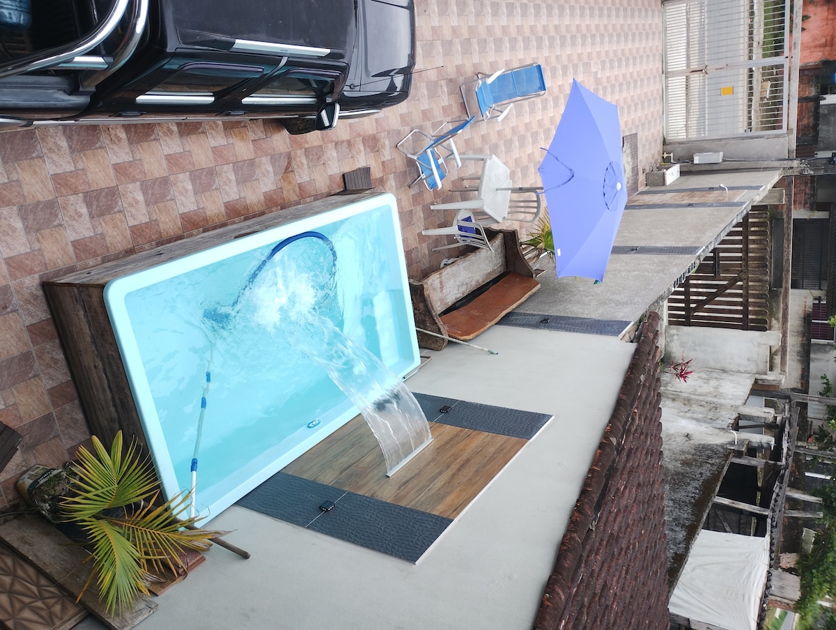 A rectangular pool is visible surrounded by a paved area, with a small waterfall feature flowing into it. A blue umbrella and two chairs are set nearby, while various plants add greenery to the space. The layout suggests a welcoming outdoor area for relaxation.